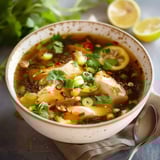 Bol fumant de bouillon avec morceaux de poulet, garni de coriandre, ciboule et piments, accompagné de tranches de citron.